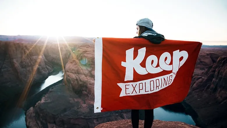 Person holding a red “Keep exploring” flag above a canyon